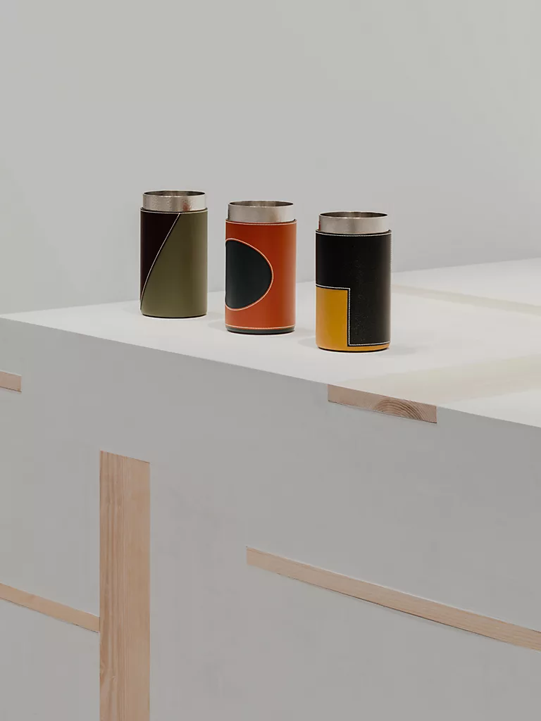 Three leather-wrapped metal tumblers in olive, orange with a black circle, and black with a yellow panel, lined up on a white display surface.