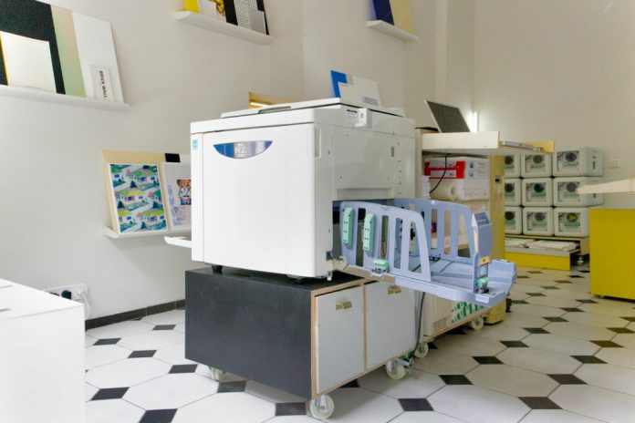 Large white printer/copier on a rolling cart with extended paper tray in a bright print shop workspace.