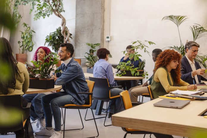 Group of people sitting at round tables in a bright plant-filled room, chatting and working on laptops and papers.