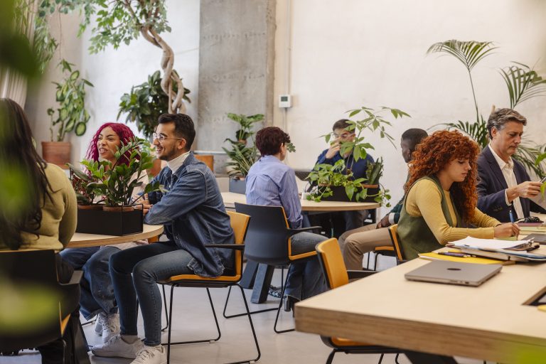 Group of people sitting at round tables in a bright plant-filled room, chatting and working on laptops and papers.