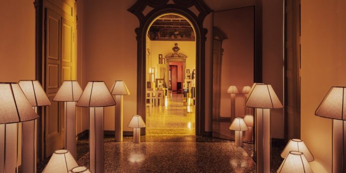Dimly lit hallway lined with numerous floor lamps, shadows softly glowing toward an ornate doorway at the far end.