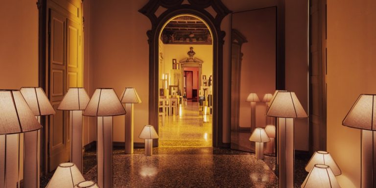 Dimly lit hallway lined with numerous floor lamps, shadows softly glowing toward an ornate doorway at the far end.