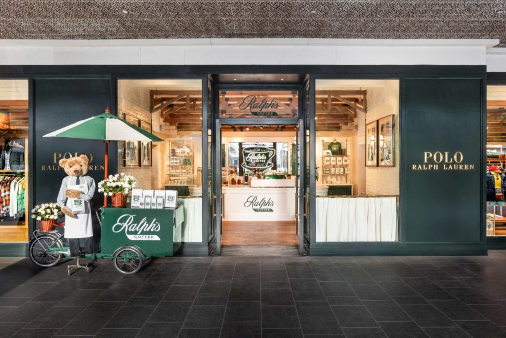 Teddy bear wearing a barista apron stands beside a green Ralph's Coffee cart with a white umbrella outside a Polo Ralph Lauren store entry.