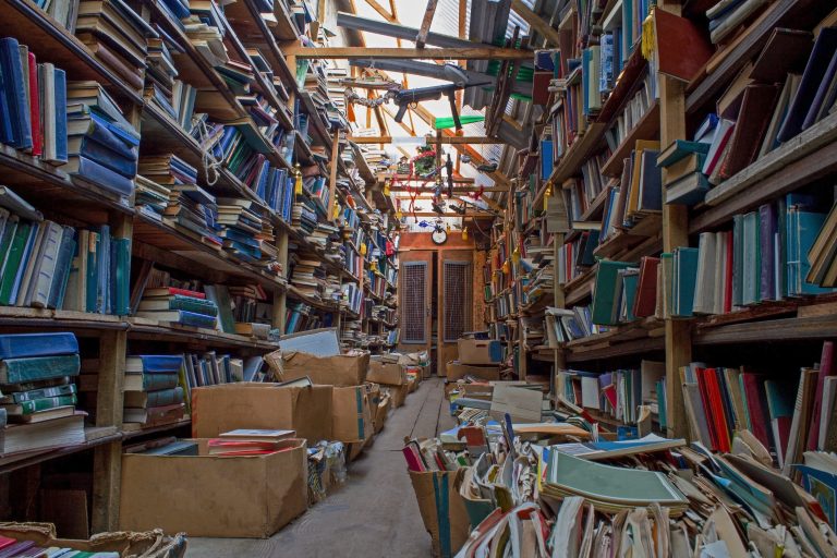 A narrow, cluttered library aisle with tall shelves packed with books on both sides and wooden beams overhead, creating a tunnel effect.
