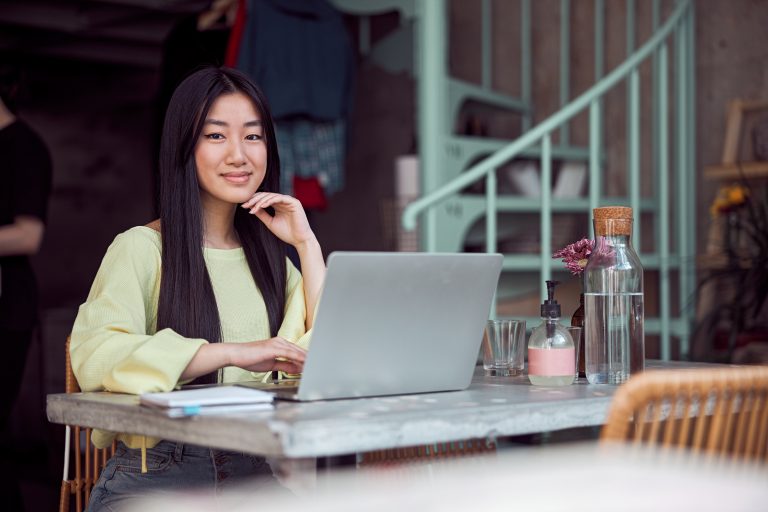 Woman with long dark hair in a yellow top sits at an outdoor café table, resting her chin on her hand while using a laptop.