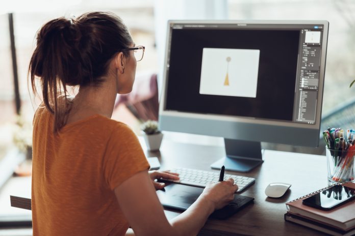 Person in an orange shirt using a graphic tablet while typing at a desk with a large monitor showing a simple design on screen