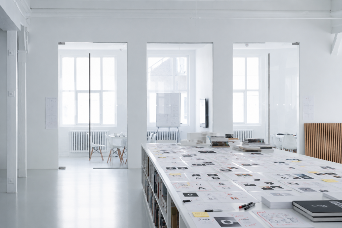 Bright white design studio with a long table covered in papers, sketches, and markers, glass-walled rooms in the background.
