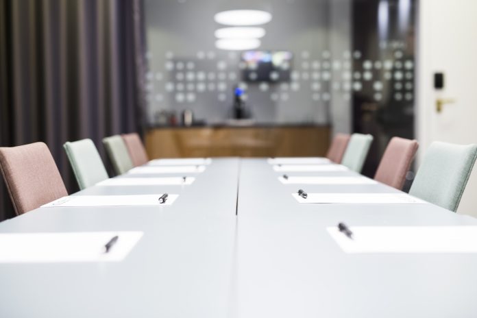 Blurred conference room with a long meeting table, papers and pens at each seat, and pastel chairs nearby.