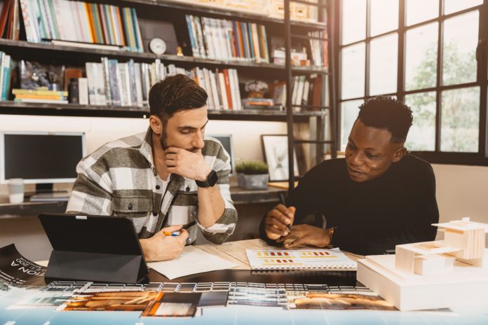 Two people collaborate at a desk, reviewing color swatches and documents in a bright office with bookshelves behind them.